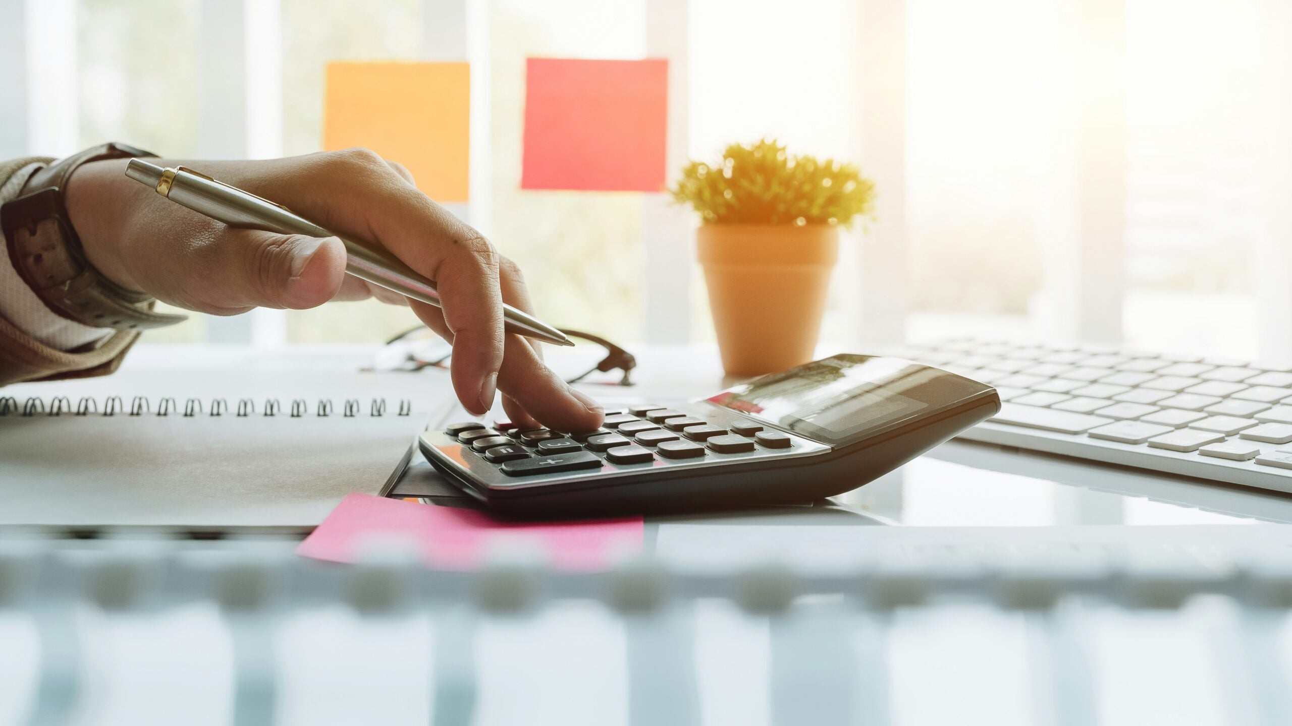 A hand using a calculator on a desk with a pen, notebook, keyboard, and small potted plant. Bright lighting and sticky notes are in the background.