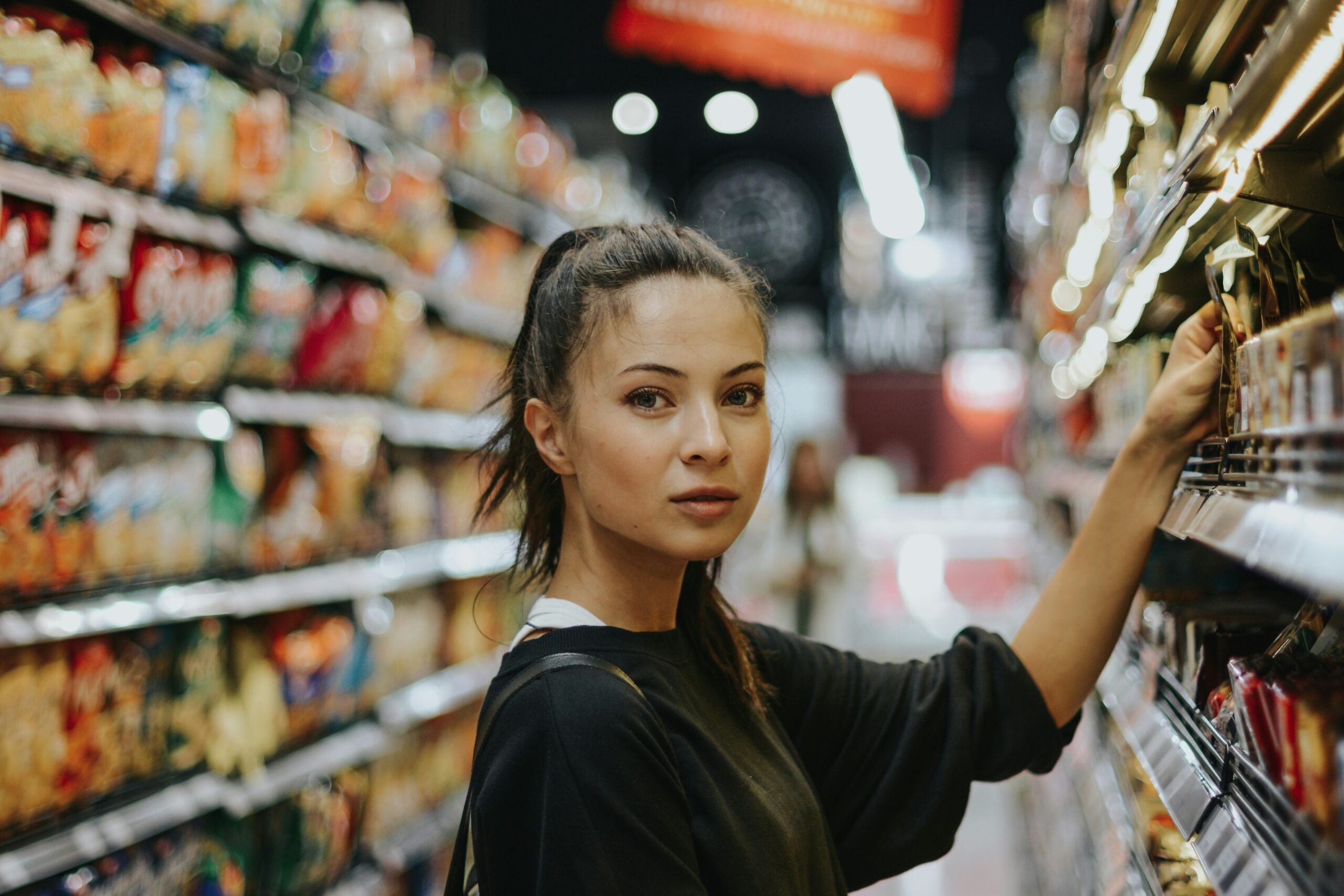 A person with long hair in a ponytail is shopping in a grocery store aisle filled with colorful packaged goods.