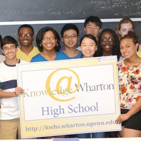 A group of young people holding a sign that reads "Knowledge @ Wharton High School" with the website URL kw.wharton.upenn.edu, in front of a chalkboard.