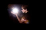 A person wearing glasses holds a glowing, bright light source in their hands, set against a dark background.