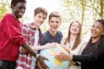 A group of five smiling young people outdoors, holding a globe together, symbolizing unity and global connection.