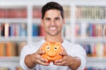 A person holds an orange piggy bank with a smiling expression, in front of a blurred background of bookshelves. This image likely represents saving money or financial planning.