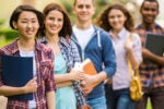 A group of five young adults, casually dressed, standing in a line outdoors, each holding books or folders. They appear to be students.