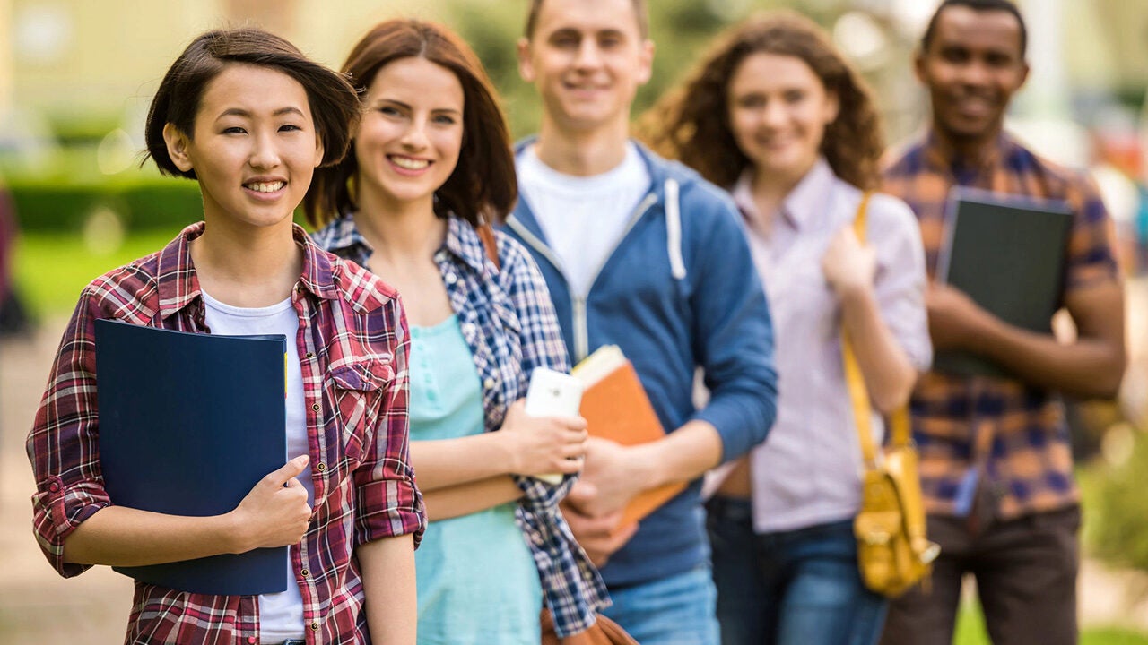 A group of five young adults, casually dressed, standing in a line outdoors, each holding books or folders. They appear to be students.