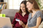Two people sitting on a couch shopping online with a laptop, holding a credit card, and smiling.