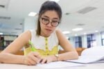 A person with glasses is studying in a library, writing notes on paper next to an open book.