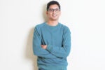 A person wearing glasses and a green sweater is leaning against a wall with arms crossed, smiling.