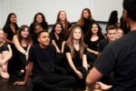A group of people sit on the floor in a dance studio, wearing black clothes, watching a person in the foreground. They appear engaged and attentive.