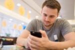 A person in a casual gray t-shirt is sitting at a table, looking at a smartphone. The background appears to be a bright, modern indoor setting.