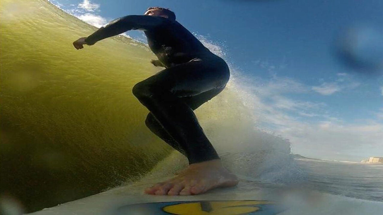 A surfer in a wetsuit riding a wave, captured from a low-angle perspective on the surfboard, with a clear blue sky in the background.