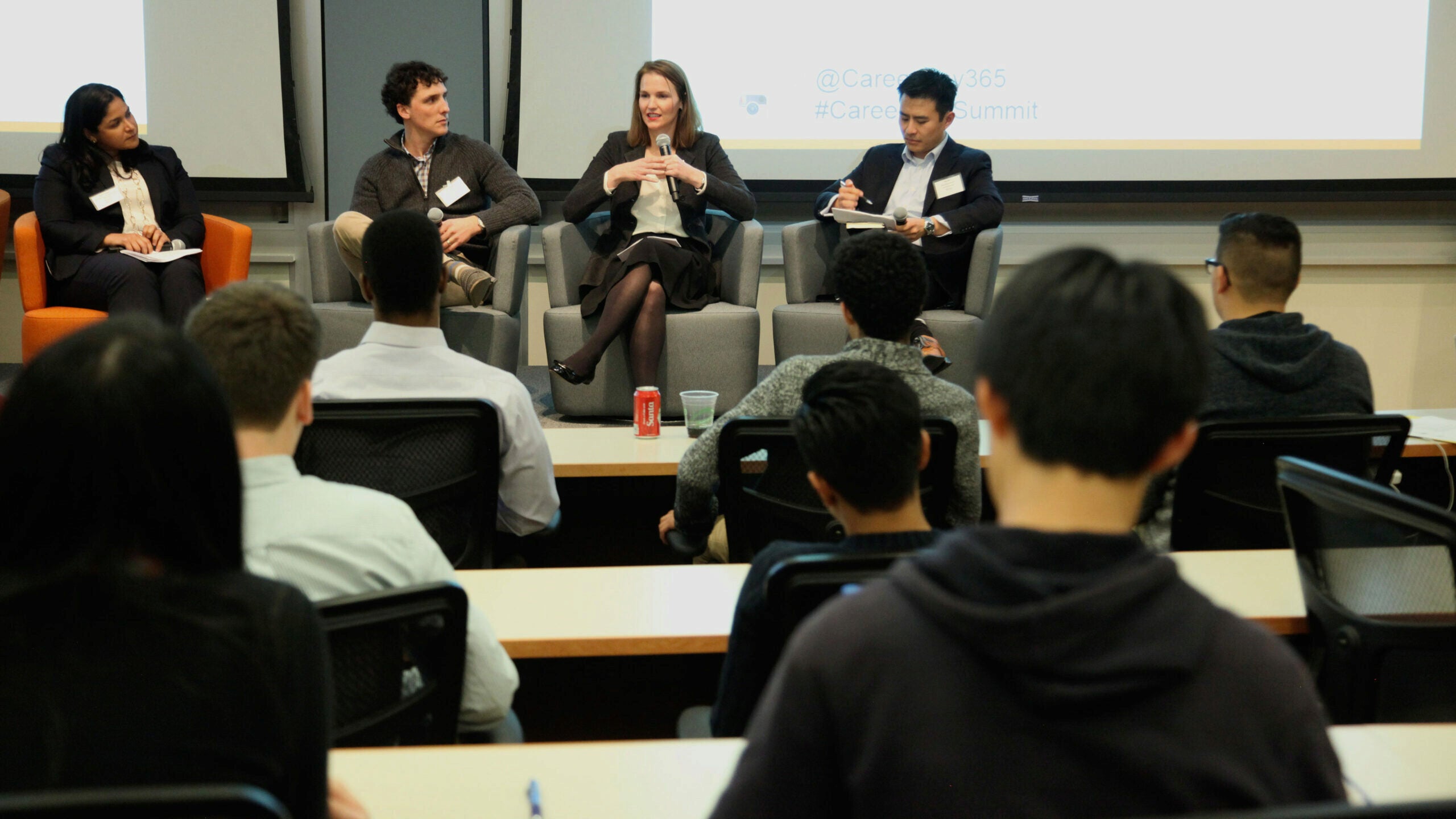 A panel discussion with four people seated on stage, engaging with an audience in a conference setting. Audience members are seated at tables, facing the panelists.