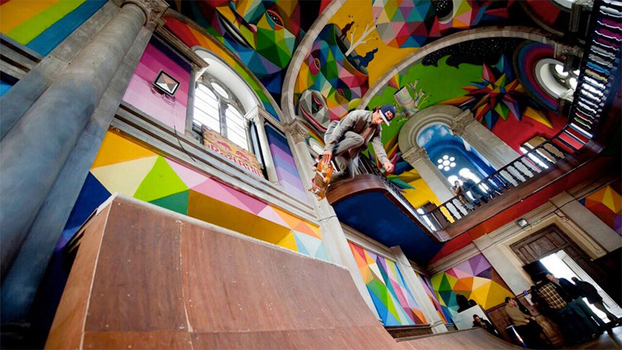 A skateboarder performs a trick inside a vibrant, graffiti-covered church interior with geometric and colorful designs.