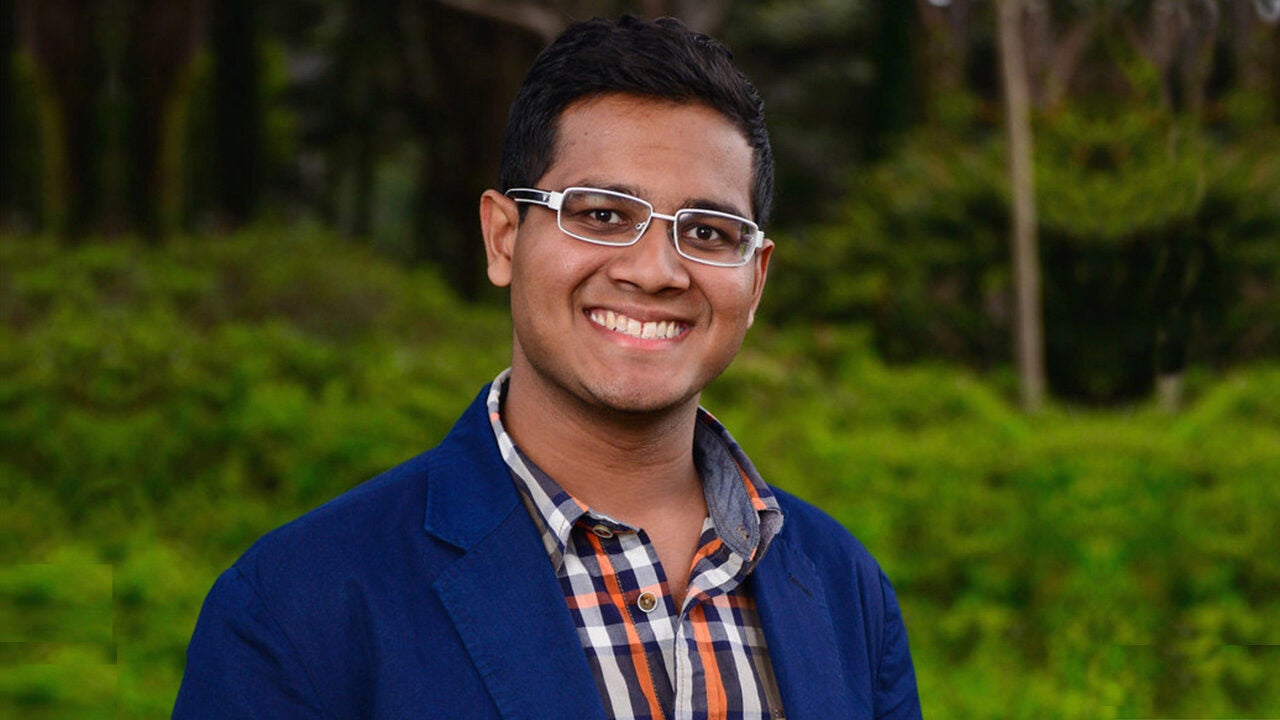 A person wearing glasses and a blue blazer, smiling outdoors with greenery in the background.