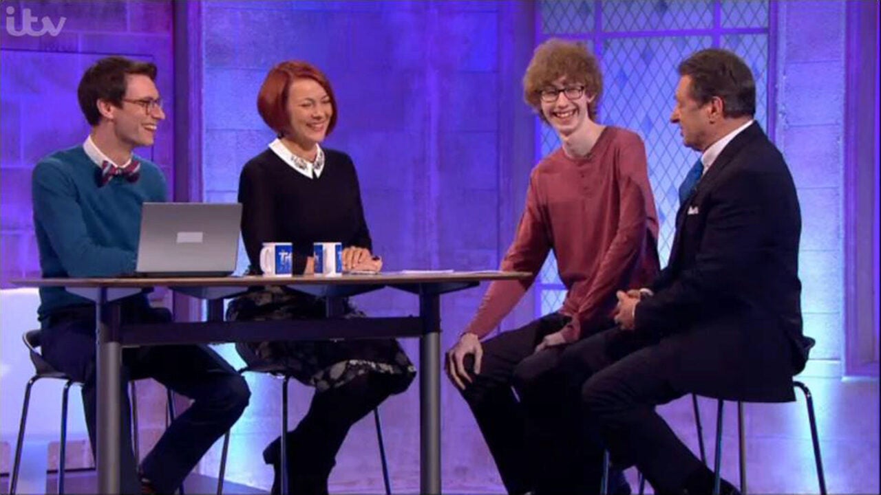 Four people are sitting together indoors on a television set, smiling and engaged in conversation. There is a laptop and mugs on the table.