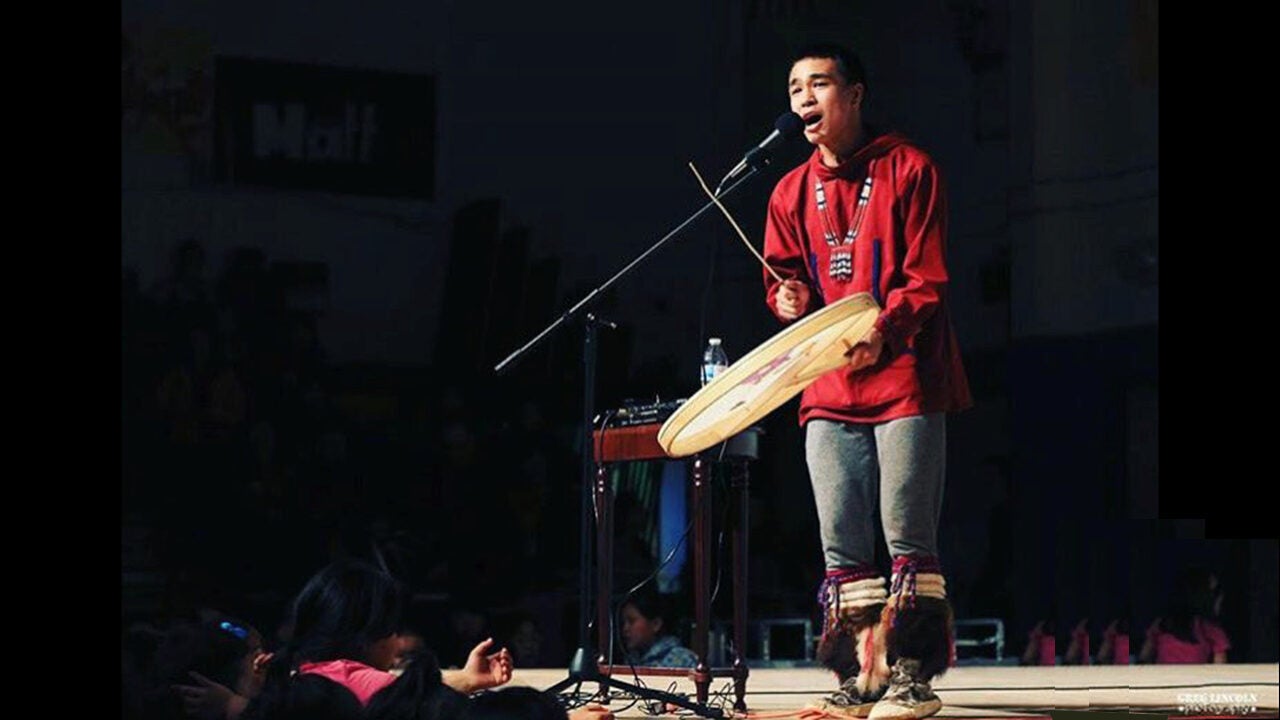 Person in traditional attire performing on stage, holding a drum and singing into a microphone. Audience visible in the background.