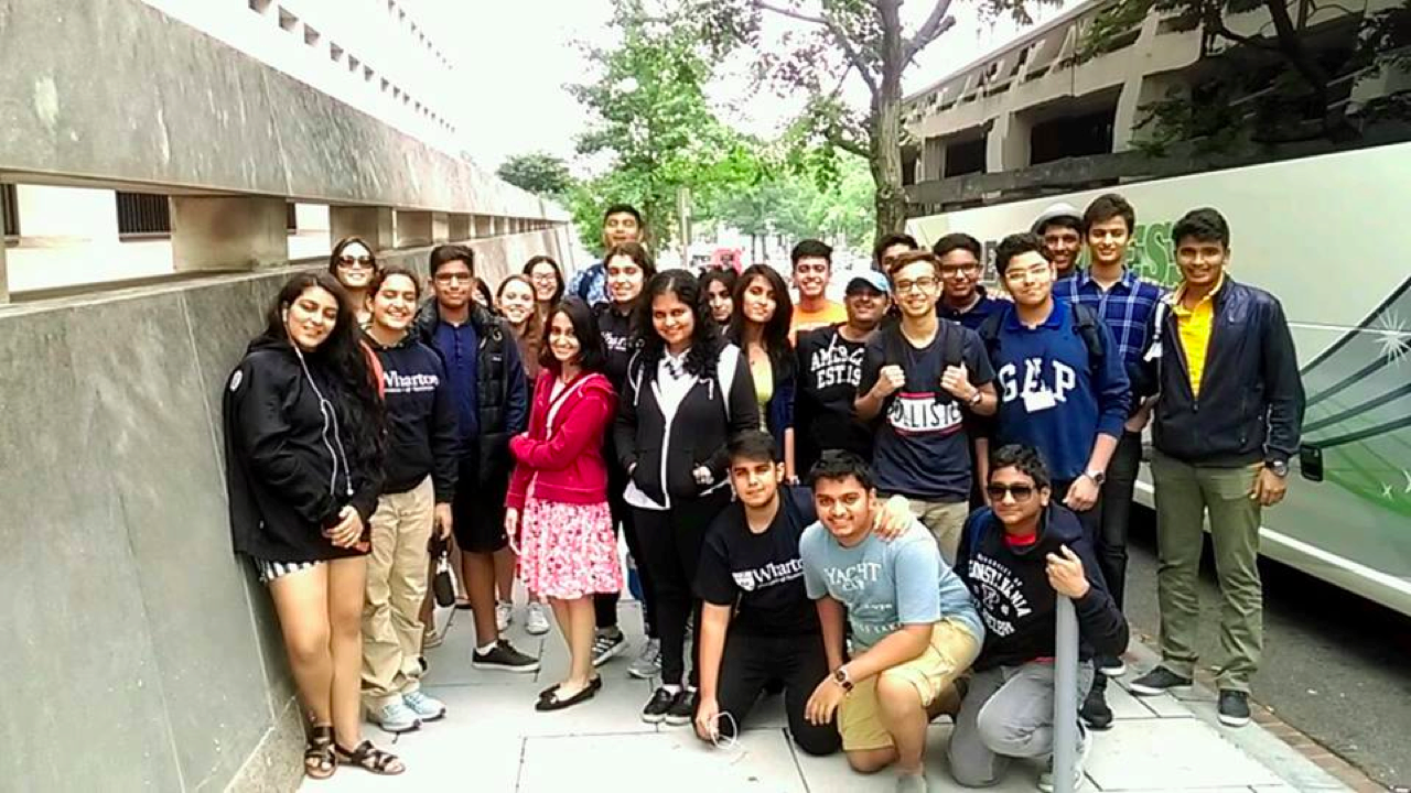 A group of people standing together outdoors; some are smiling. They are next to a building and a parked bus, with trees in the background.