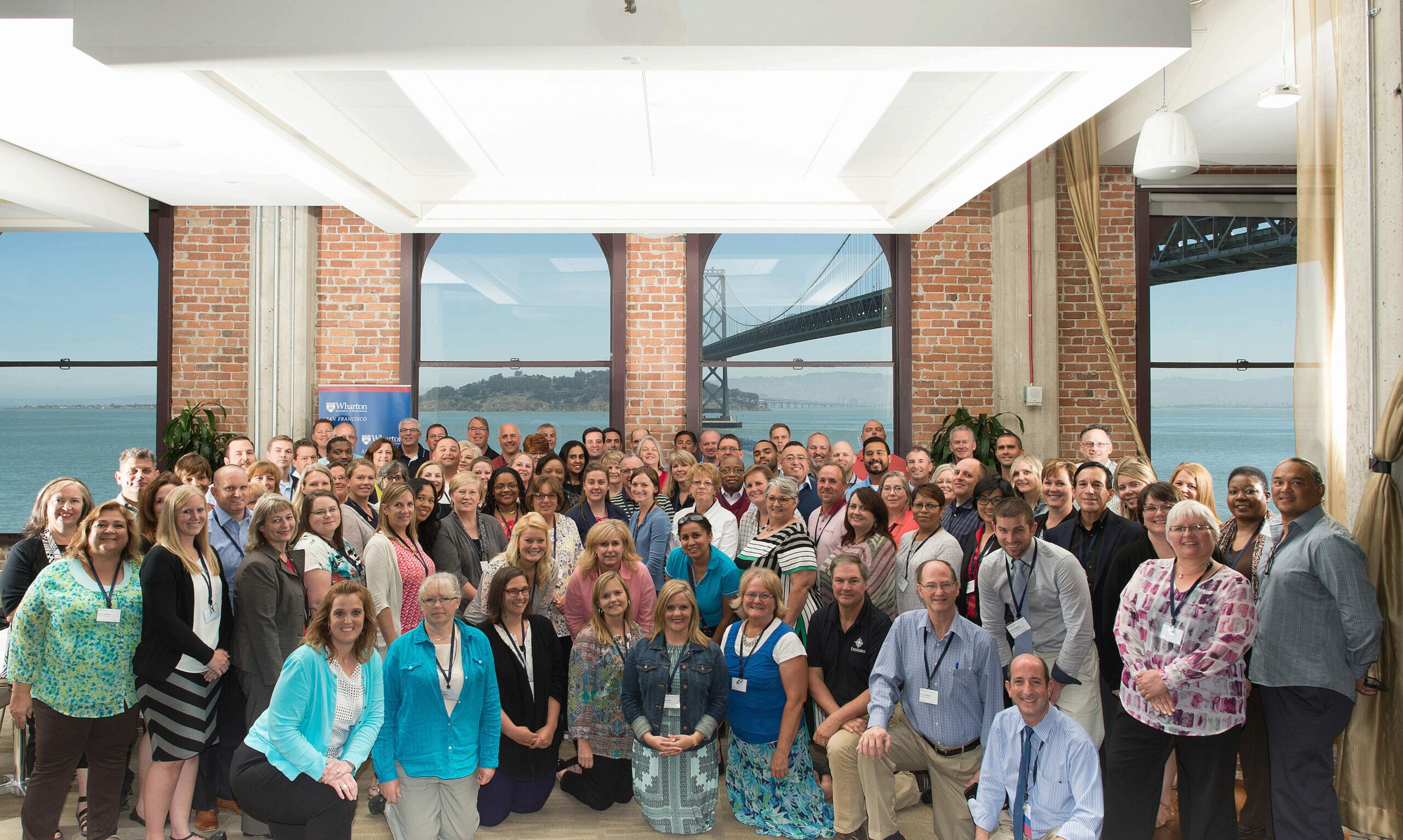 A large group of people are gathered in a well-lit room with brick walls and large windows showcasing a view of a bay and bridge. They are posing for a group photo, many wearing name tags.