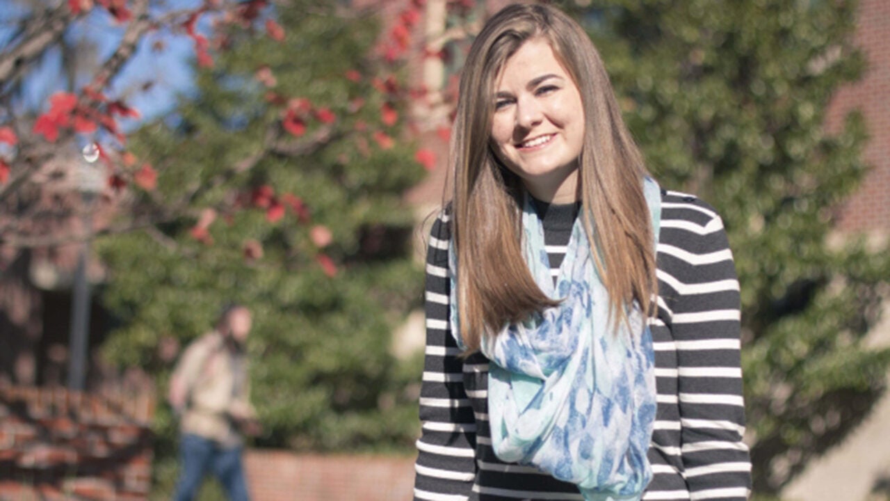 A person with long hair is smiling outdoors, wearing a black-and-white striped top and a light blue scarf, with trees and red flowers in the background.