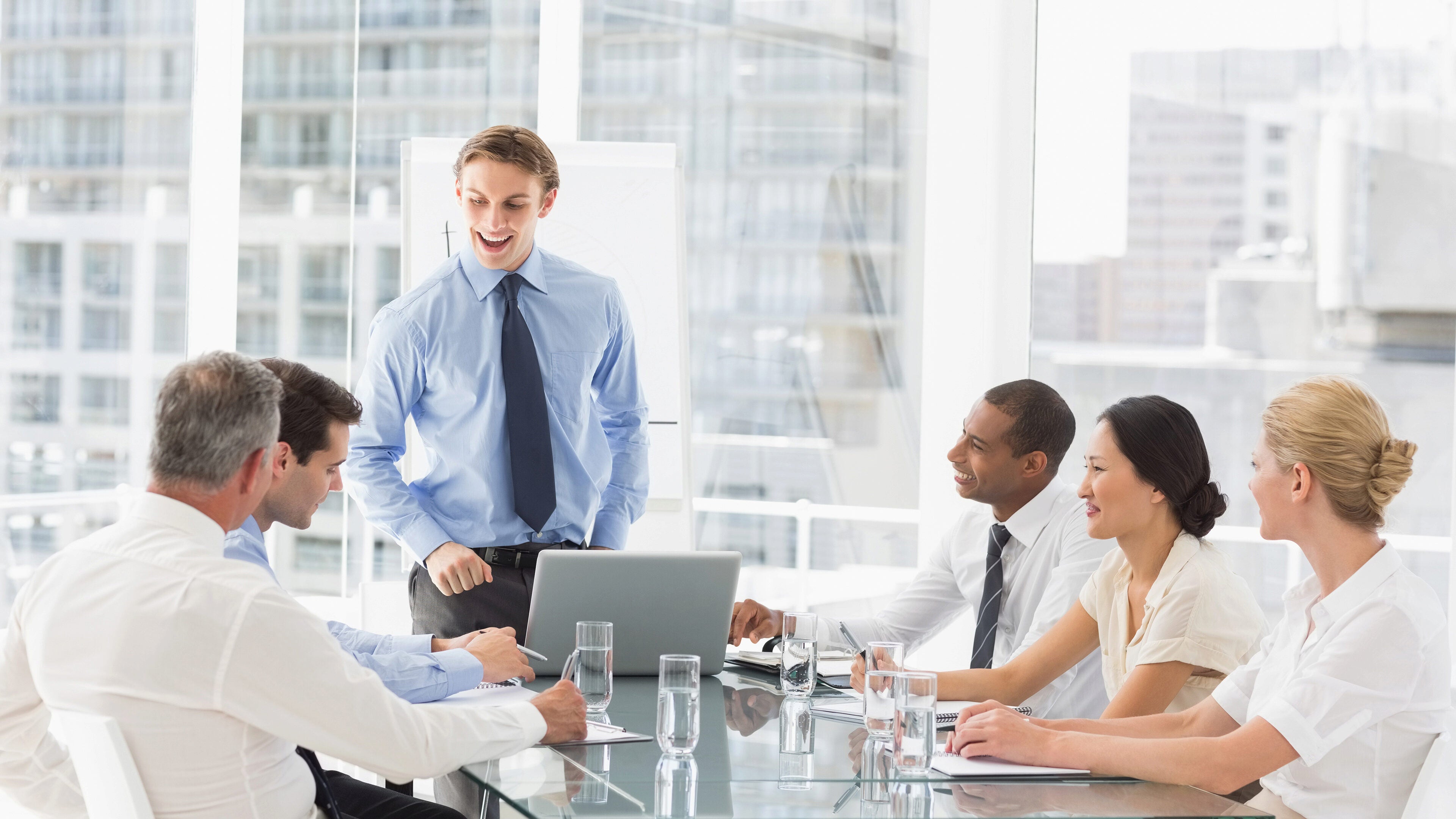 A group of people in business attire sitting around a conference table, with one person standing and speaking, suggesting a meeting or discussion.