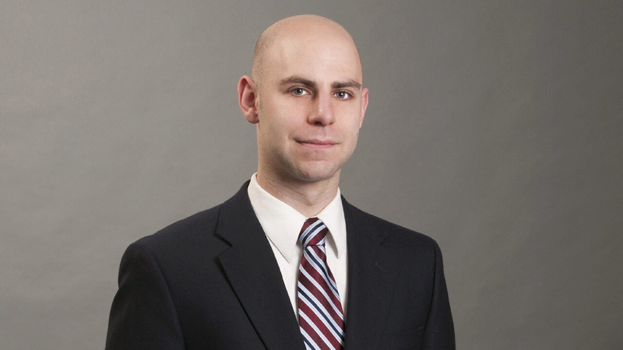 A professional headshot of a person wearing a suit and striped tie, set against a plain background.