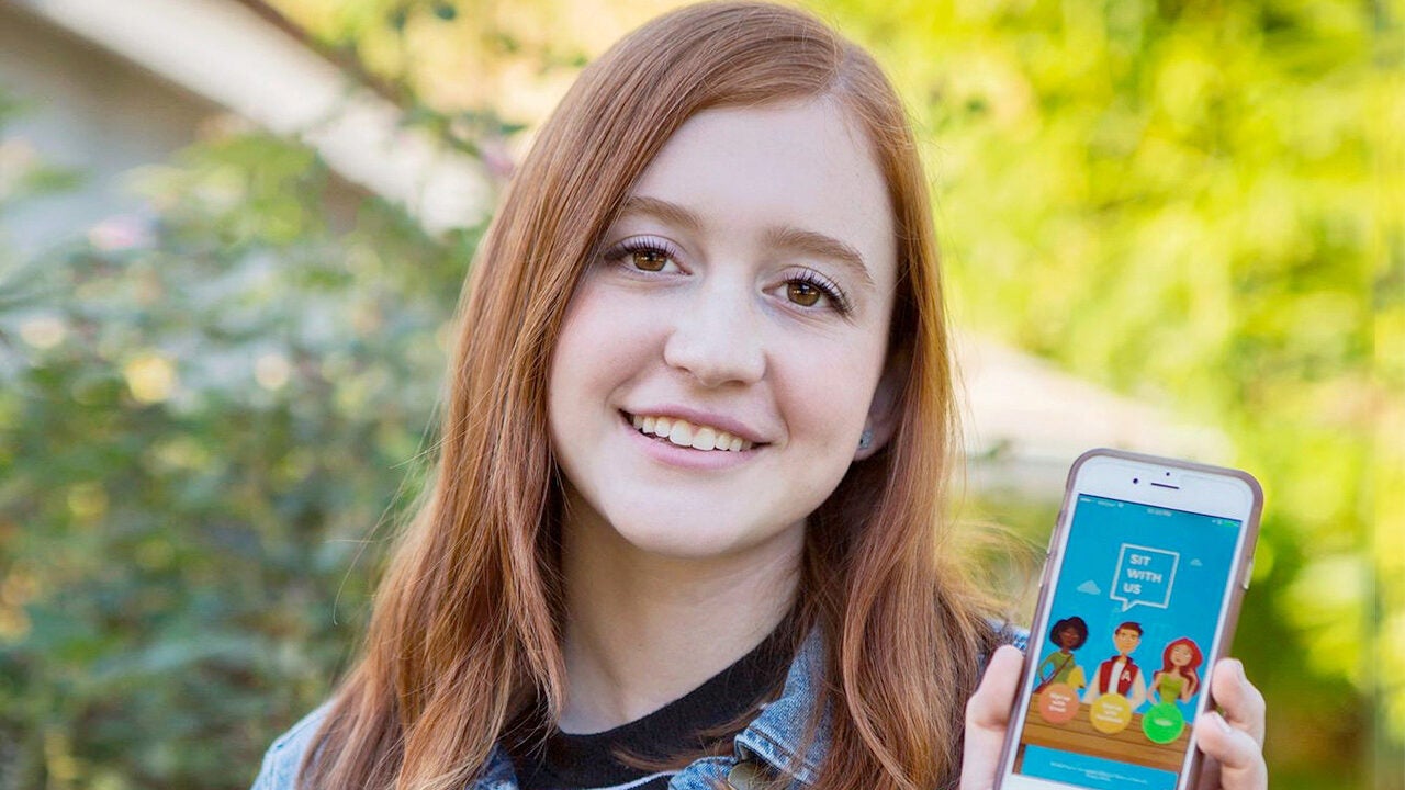 A person smiling and holding a smartphone displaying an interface with colorful cartoon characters and the text "Sit With Us."