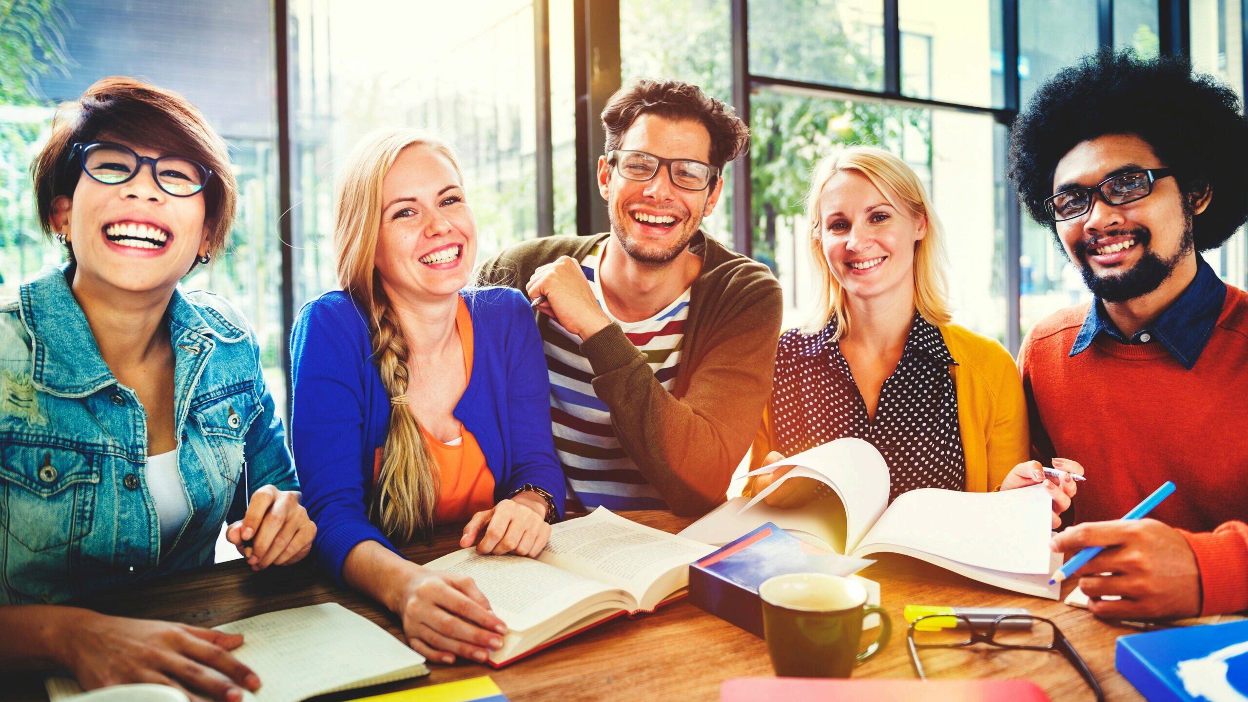 A diverse group of five people sitting together at a table, smiling and studying with open books and notebooks, in a bright, casual setting.