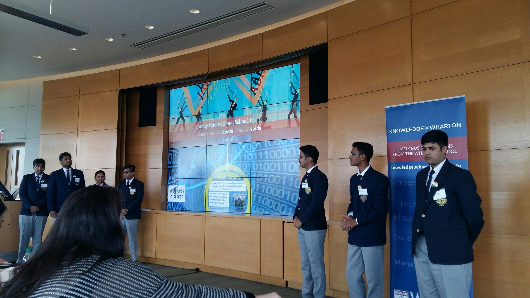 A group of students in formal attire stand facing a screen displaying the names "Amity International School" and text about a project, with a banner reading "Knowledge @ Wharton" beside them.