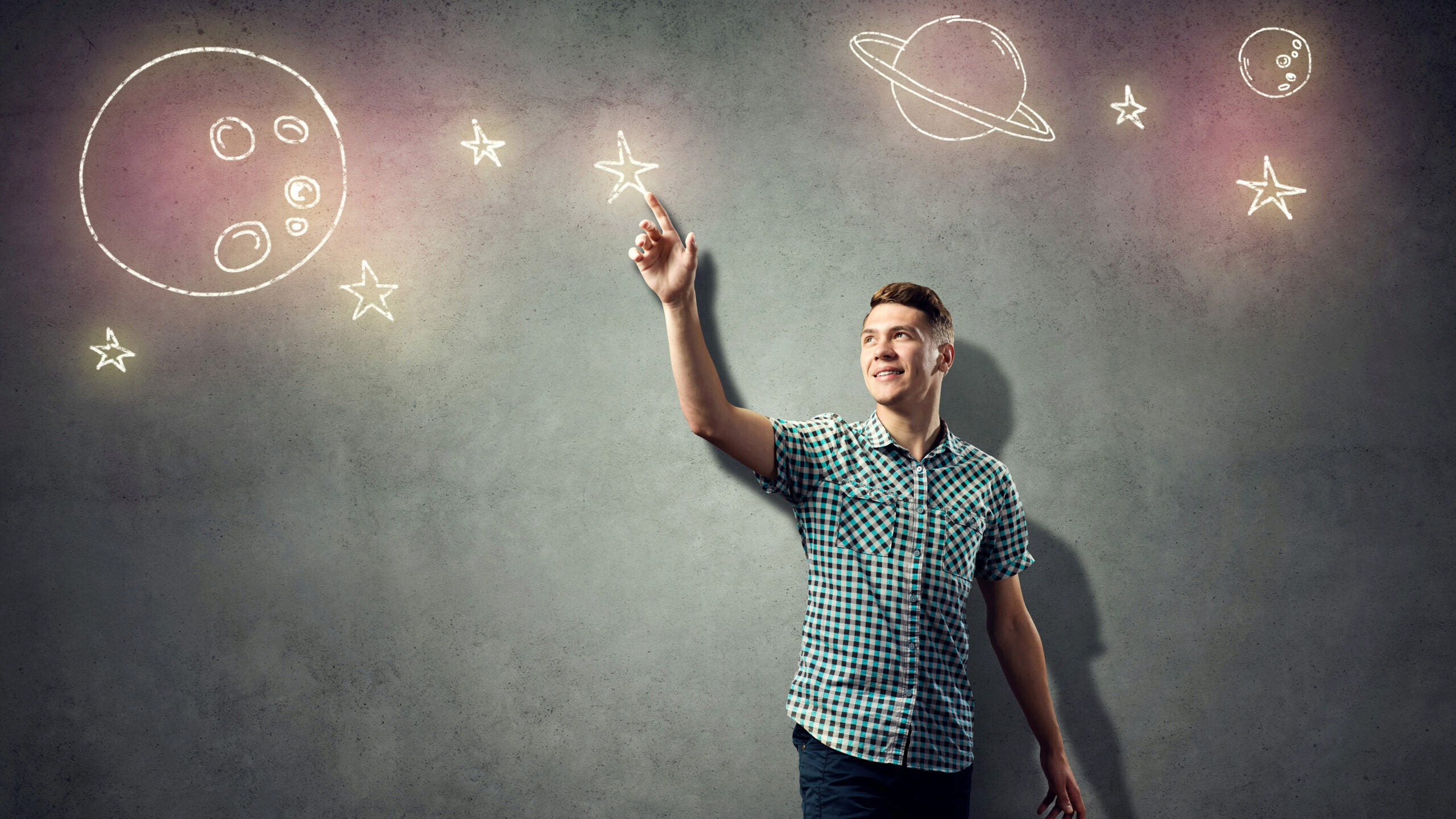 A person in a checkered shirt points at chalk-like drawings of planets and stars on a gray wall, suggesting imagination and creativity.