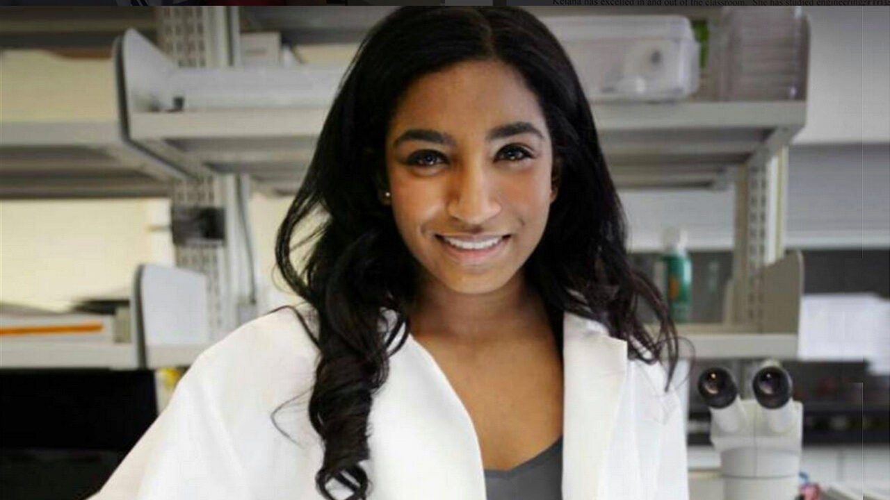 A person wearing a lab coat is smiling in a laboratory setting with shelves and a microscope in the background.