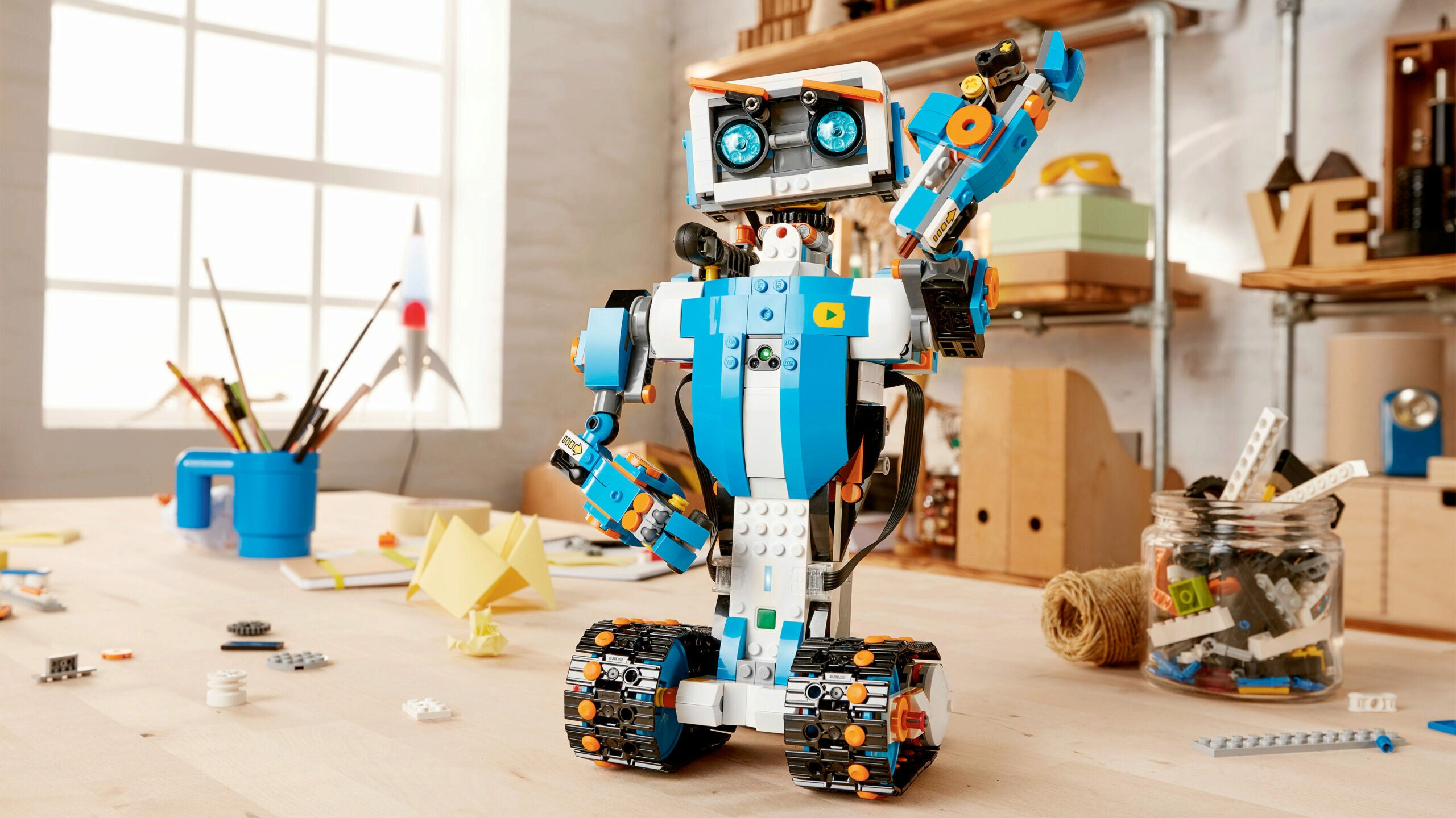 A colorful LEGO robot stands on a wooden table in a workshop setting. It has tracked wheels and a mechanical arm, with tools and LEGO pieces scattered around.