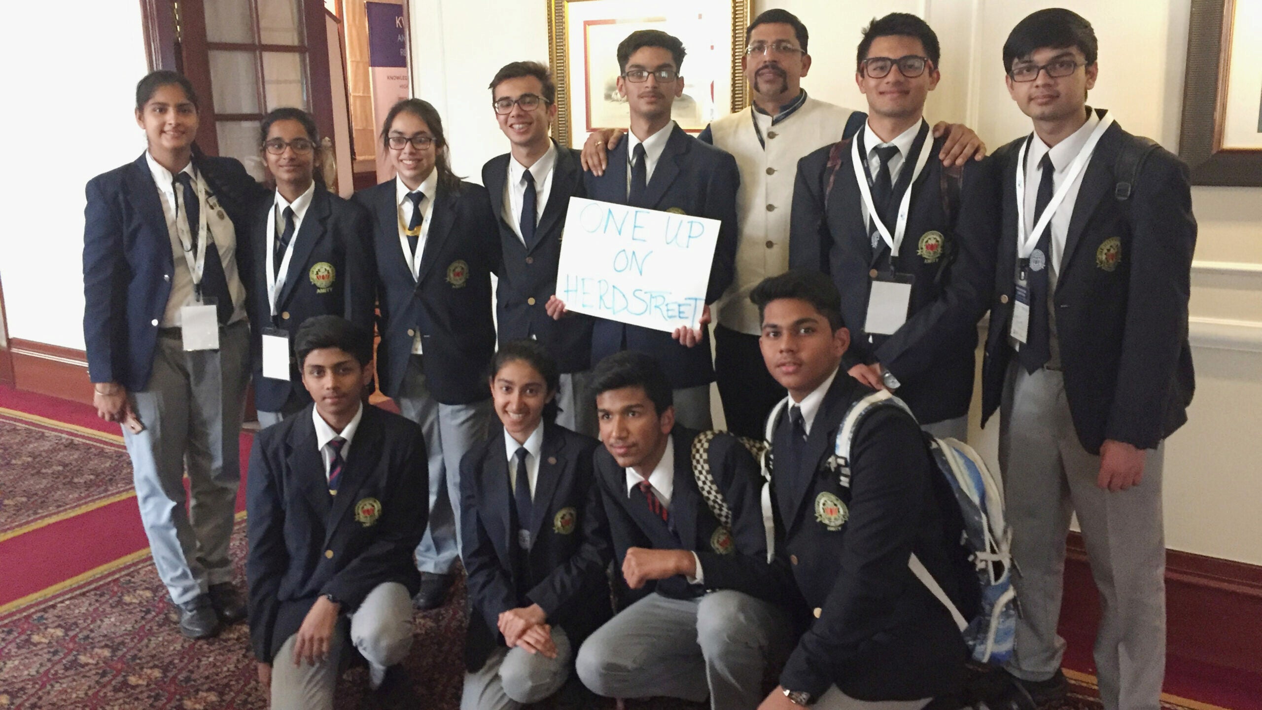 A group of students in school uniforms pose indoors with a sign reading "One Up on Herdstreet." They appear to be part of an academic or formal event.