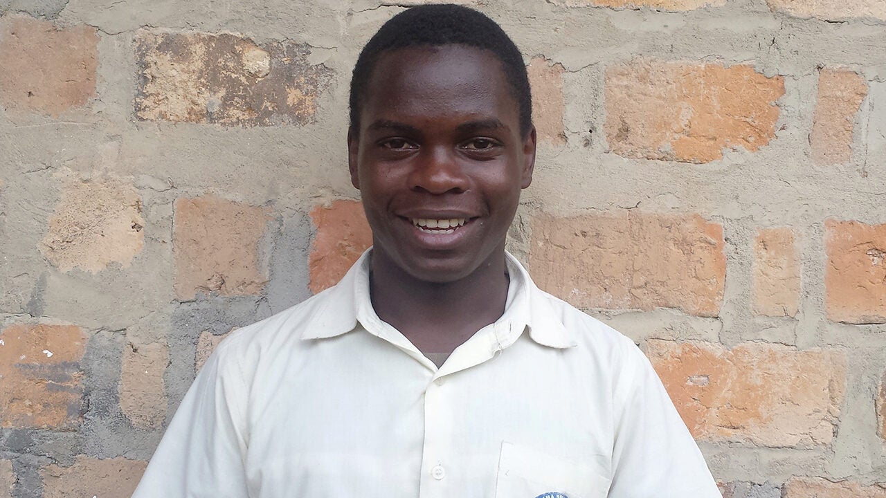 A smiling person in a white shirt stands against a textured brick wall.