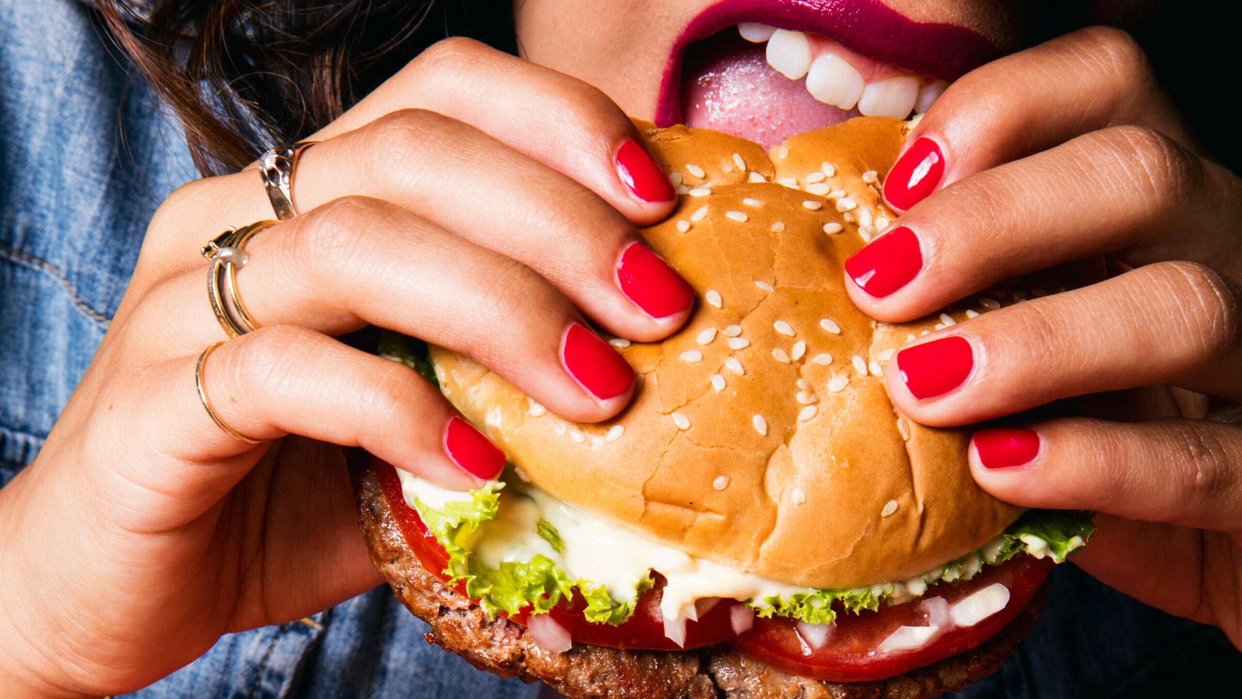 A person with red nail polish and rings holds a large sesame seed hamburger, about to take a bite. The hamburger contains lettuce, tomato, and sauce.