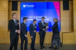 A person hands a trophy to a group of young individuals wearing suits. A large screen in the background shows text related to the 2017 KWHS Investment Competition at the Wharton School.