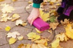 A hand picking up a pink wallet from a leaf-covered stone path, with a person wearing purple and black shoes nearby, suggesting an autumn scene.