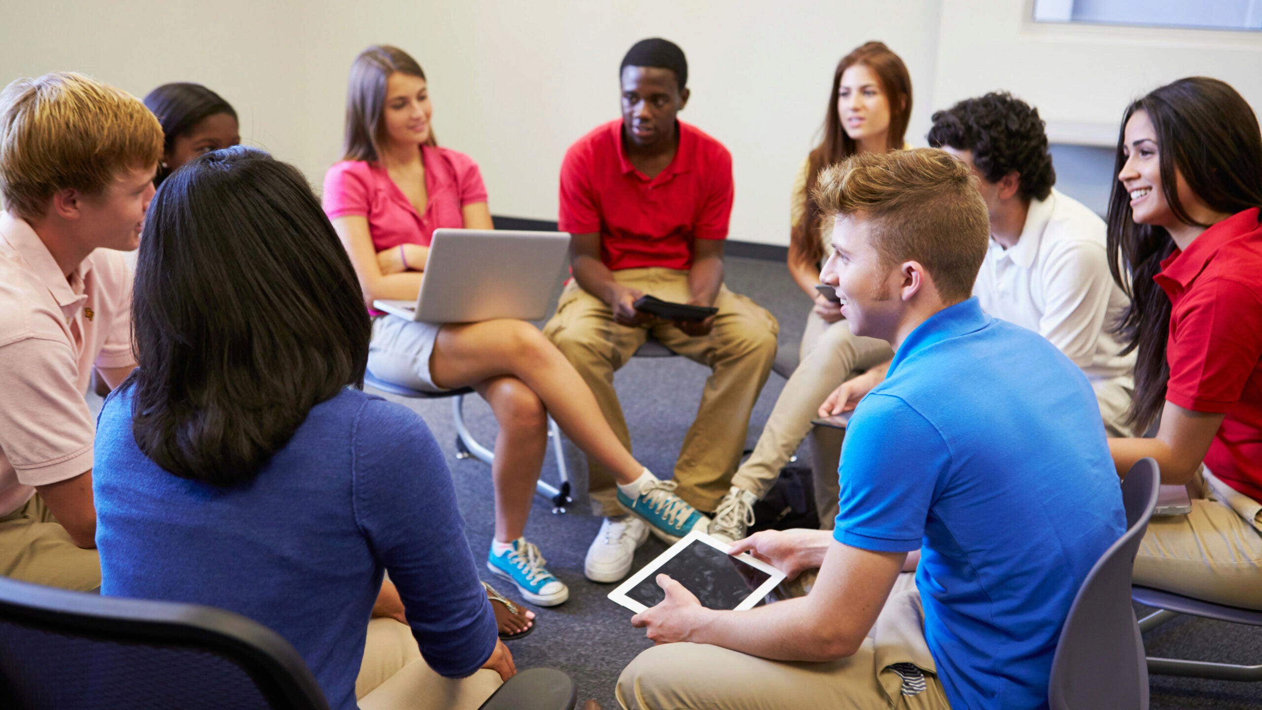 A group of young people sit in a circle, engaged in discussion. Some hold electronic devices like tablets and laptops. They are in a casual setting, possibly a classroom or meeting room.