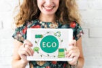 A person holding a tablet displaying eco-friendly symbols, including a light bulb with leaves, a battery, and a recycling symbol, with the word "ECO" in the center. The background is a white brick wall.