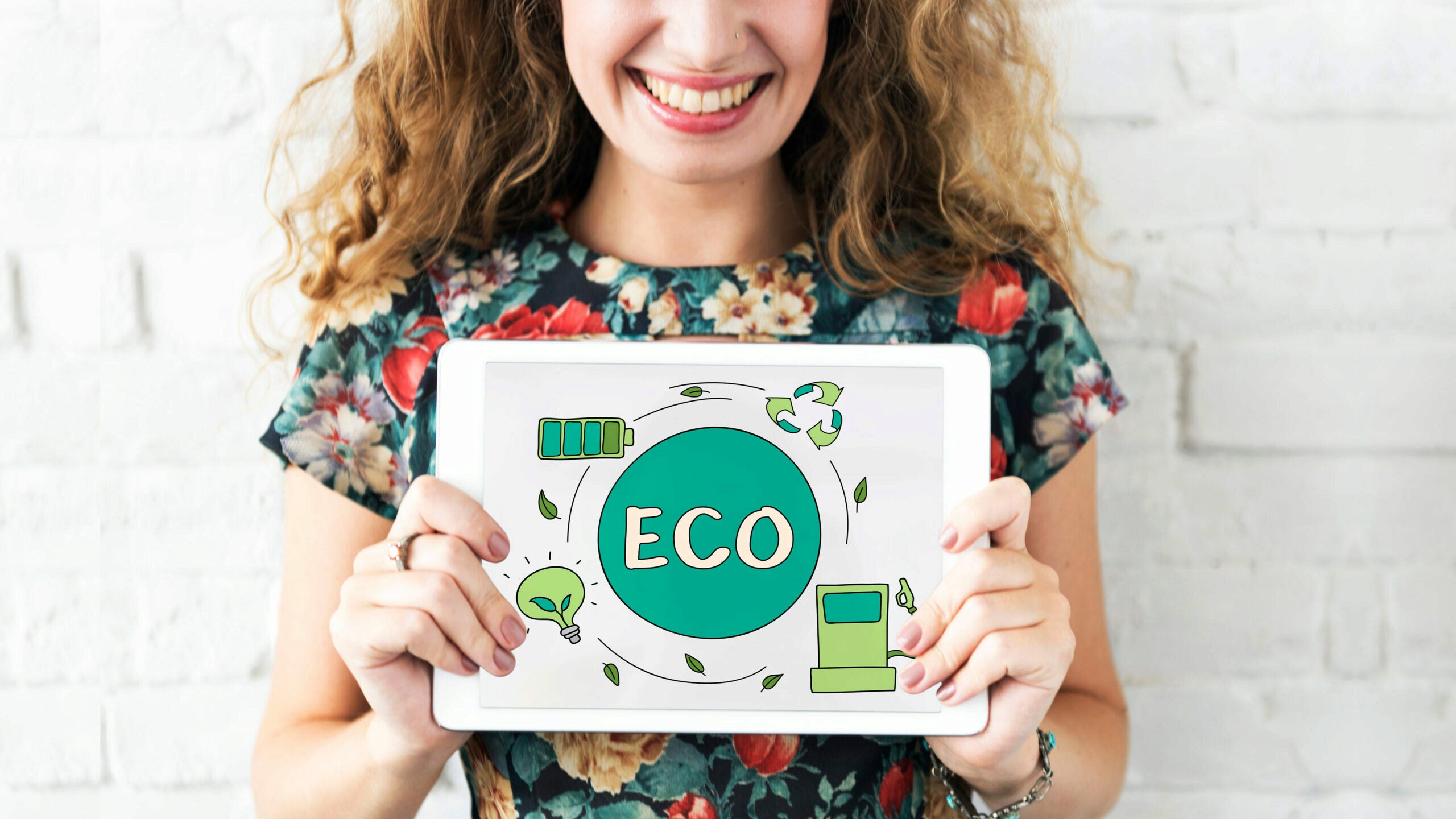 A person holding a tablet displaying eco-friendly symbols, including a light bulb with leaves, a battery, and a recycling symbol, with the word "ECO" in the center. The background is a white brick wall.