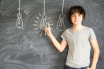 A person in a gray t-shirt stands in front of a blackboard with chalk drawings of three light bulbs. One bulb is illuminated, and the person is pointing at it, smiling.