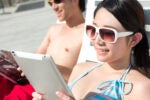 Two people relaxing on lounge chairs, wearing sunglasses. One is reading a magazine, and the other is using a tablet. They appear to be enjoying a sunny day.