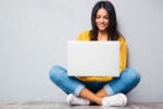 A person sitting cross-legged on the floor, using a laptop, wearing a yellow sweater and blue jeans, smiling at the screen.