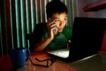 A person is sitting in a dimly lit room, using a laptop and talking on a cellphone. A blue mug and glasses are on the table nearby.