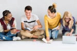 A group of four people sitting on the floor, engaged in studying or collaborating. They are smiling and have books and a laptop.