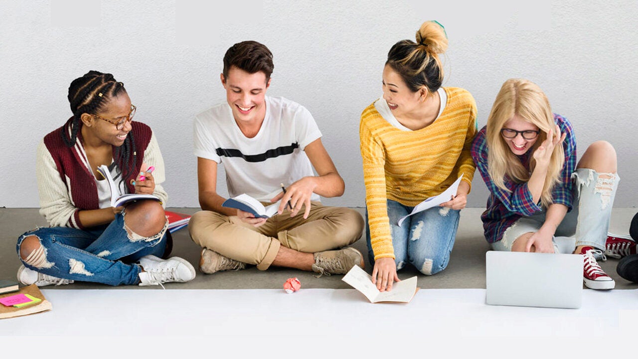 A group of four people sitting on the floor, engaged in studying or collaborating. They are smiling and have books and a laptop.