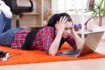 A person lying on an orange carpet in a room, looking frustrated at a laptop. Nearby are headphones and a pair of glasses.
