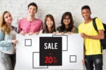 Five people smiling and holding a sign that says "SALE UP TO 20% OFF" against a white brick wall.