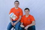 Two people wearing matching orange "Wolf''em" shirts, one seated and the other kneeling, holding a packaged roasting stick.