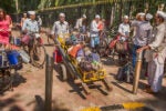 A group of dabbawalas in traditional white attire are gathered on a street with bicycles and tiffin containers. A cart is loaded with bags.
