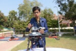 A person in a blue suit is holding a drone with a camera attached, standing in an outdoor setting with trees and a building in the background.