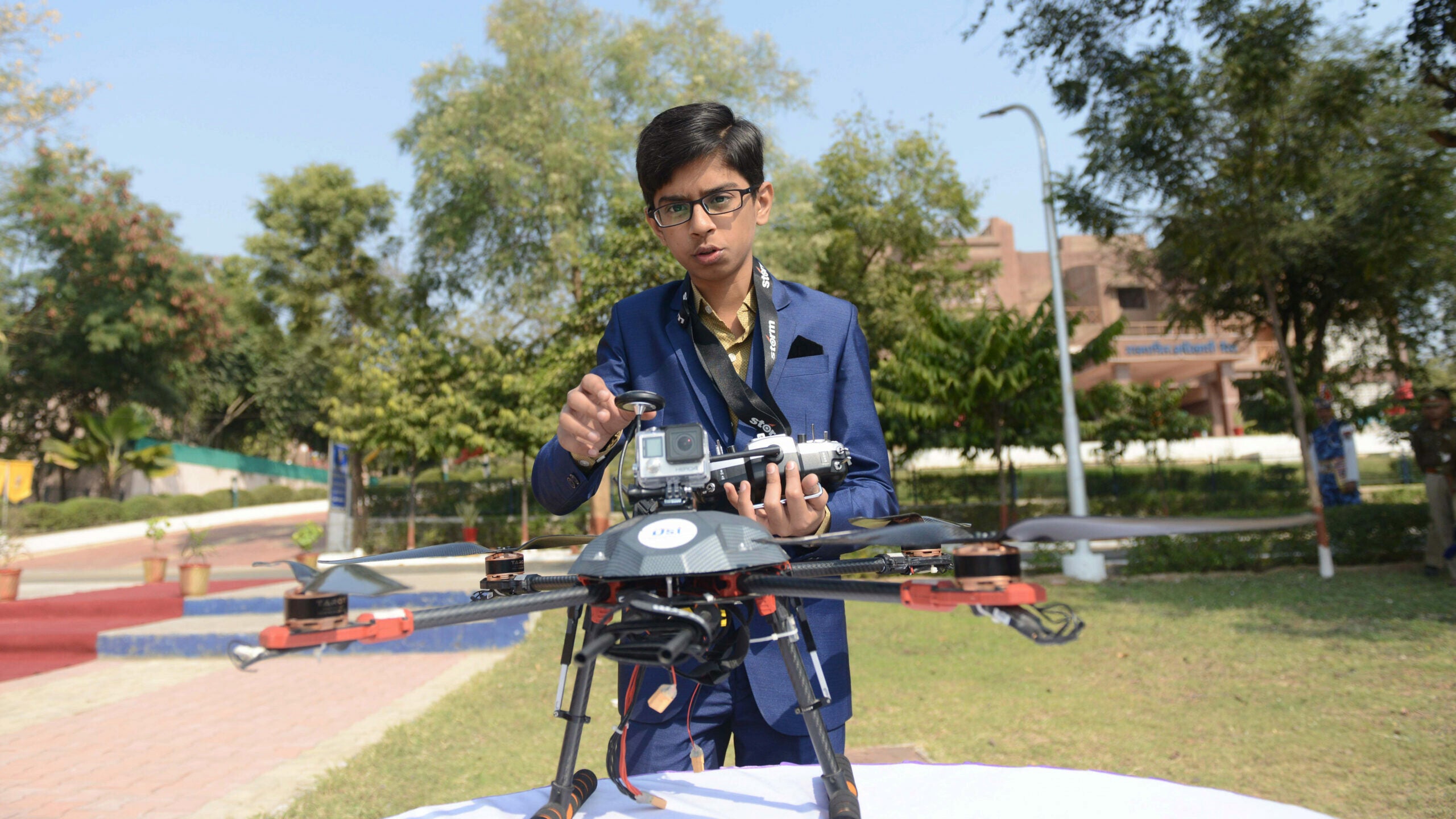 A person in a blue suit is holding a drone with a camera attached, standing in an outdoor setting with trees and a building in the background.